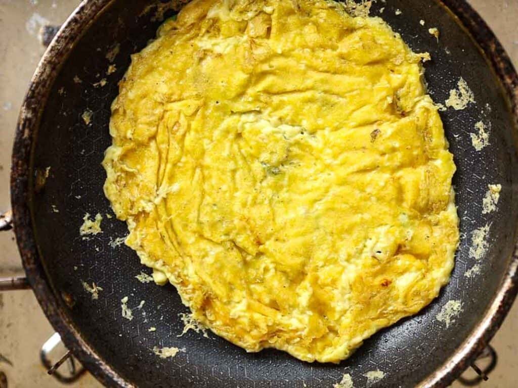 A golden-brown omelette cooking in a black frying pan. The surface of the omelette is slightly crispy with visible folds and edges. The pan is on a stovetop with bits of egg scattered around.